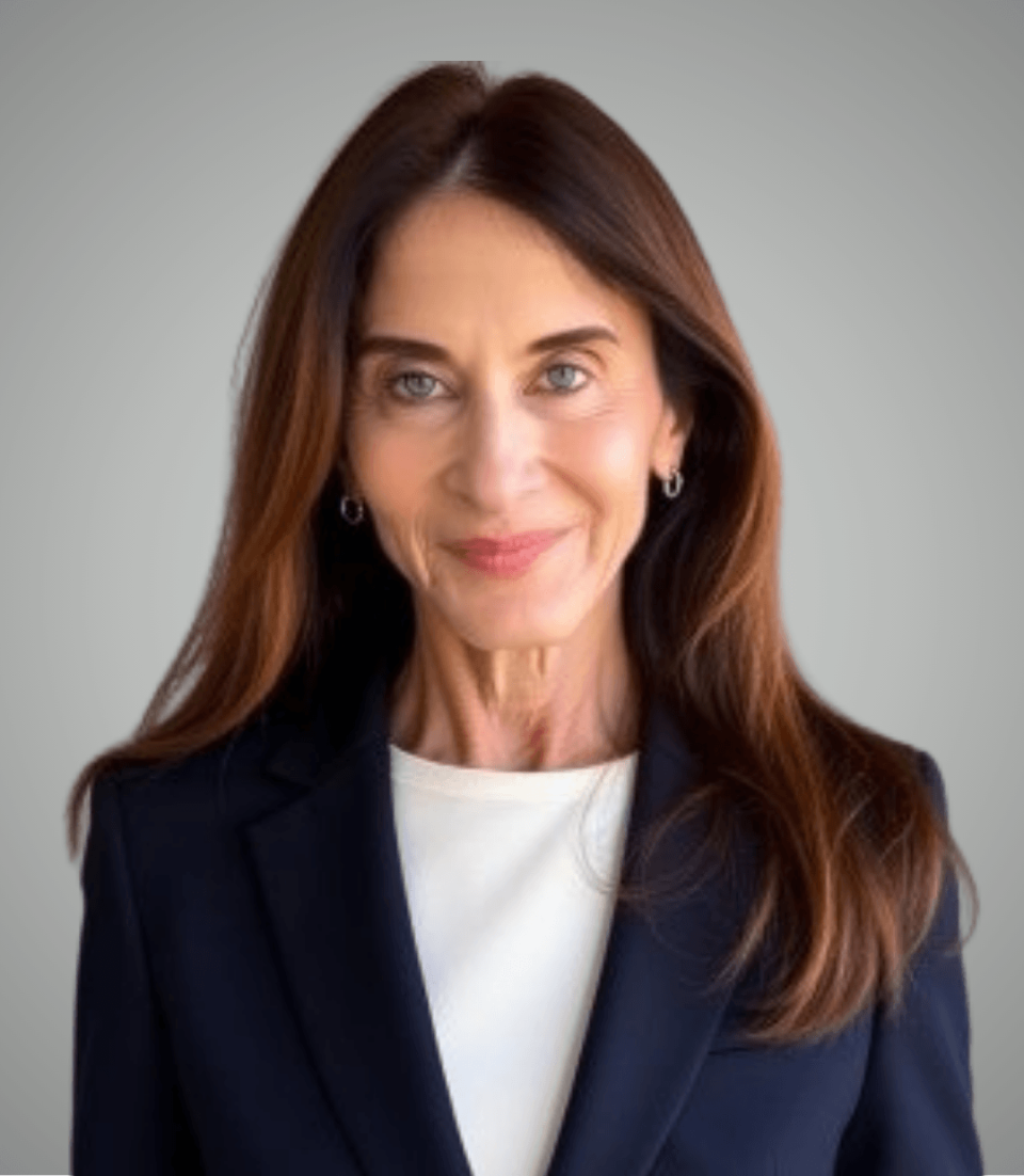 A professional woman with long brown hair, dressed in a navy blazer and white top, offers a confident, approachable smile against a plain gray background.